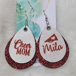 May include: A pair of red and white glitter dangle earrings. One earring says "Cheer Mom" and the other earring has a red megaphone with a heart and says "Mila".