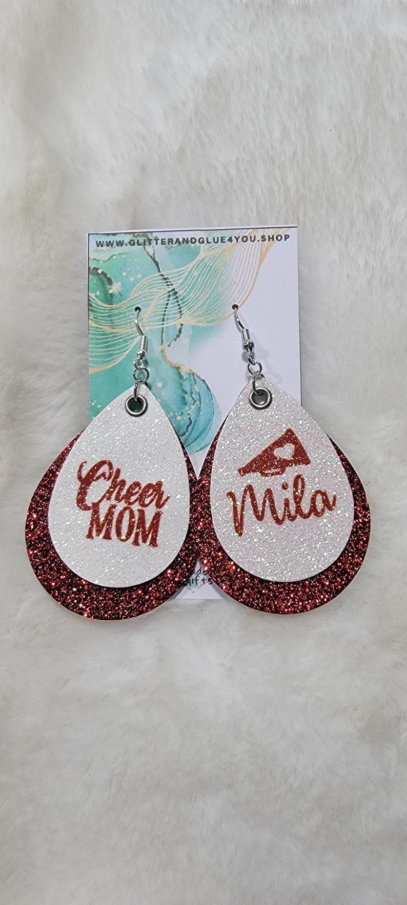 May include: A pair of red and white glitter dangle earrings. One earring says "Cheer Mom" and the other earring has a red megaphone with a heart and says "Mila".