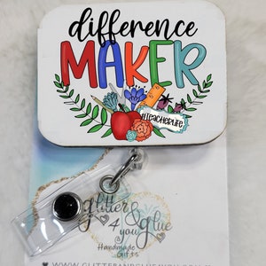 May include: A white square badge with the text "difference MAKER" in colorful letters. The badge has a floral design with a red apple, pencils, and a ruler. The badge is attached to a clear plastic retractable badge holder with a black button. The badge holder is attached to a white card with the text "Glitter & Glue 4 you Handmade Gifts www.glitterandglue4you.com" in black text.