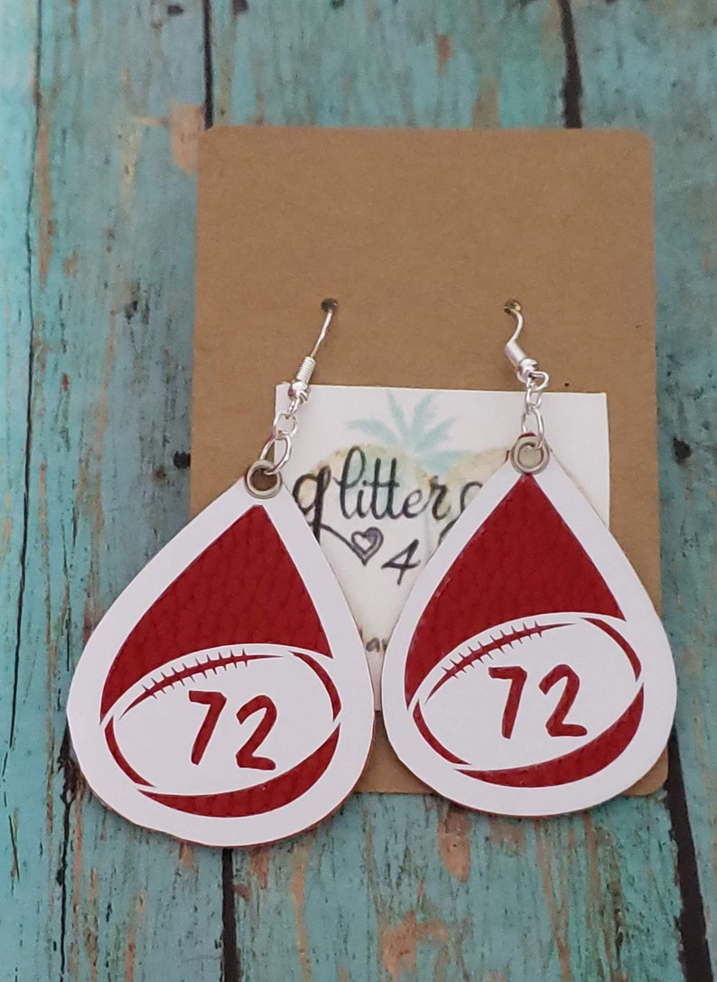 Puede incluir: Un par de pendientes de color rojo y blanco en forma de l&aacute;grima con un dise&ntilde;o de bal&oacute;n de f&uacute;tbol blanco y el n&uacute;mero "72" impreso en cada pendiente.
