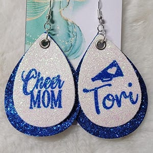 May include: A pair of white and blue glitter earrings with a blue glitter border. One earring says "Cheer Mom" and the other earring has a megaphone with a heart and says "Tori".
