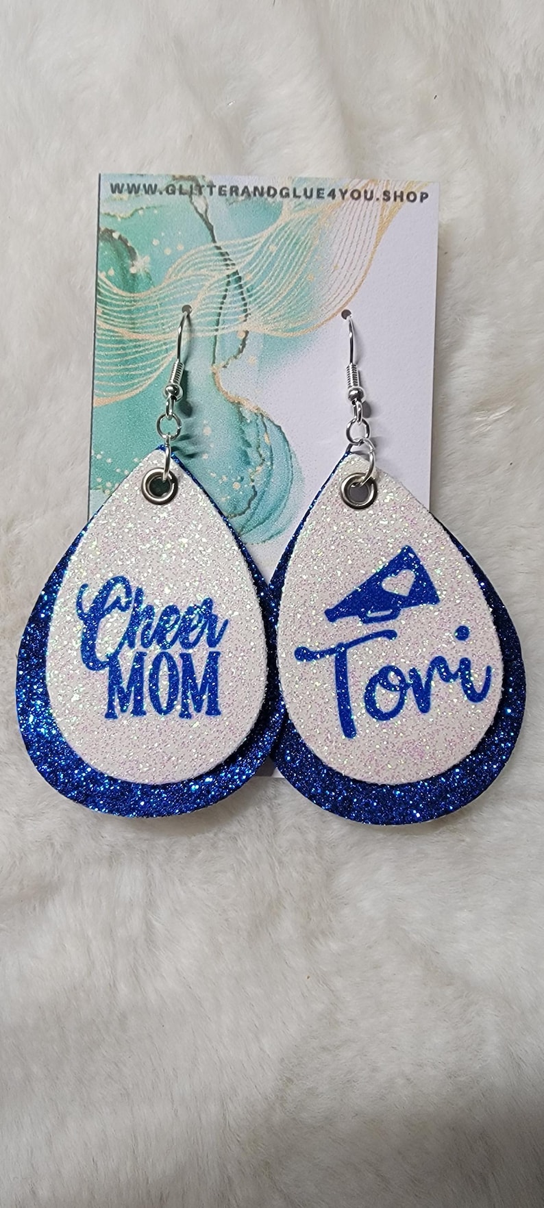 May include: A pair of white and blue glitter earrings with a blue glitter border. One earring says "Cheer Mom" and the other earring has a megaphone with a heart and says "Tori".