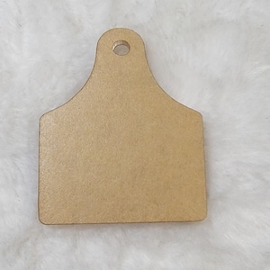 May include: A blank brown wooden tag shaped like a cow's ear with a hole at the top for hanging.