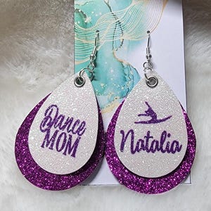 May include: A pair of dangle earrings with a white base and purple glitter. One earring says "Dance Mom" and the other earring has a silhouette of a dancer and says "Natalia".