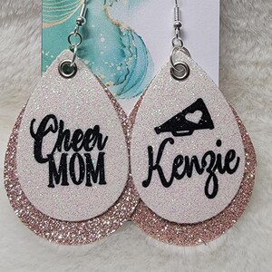 May include: A pair of white glitter earrings with a rose gold glitter trim. One earring says "Cheer Mom" and the other earring has a black megaphone and says "Kenzie".