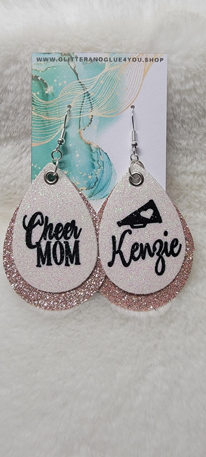 May include: A pair of white glitter earrings with a rose gold glitter trim. One earring says "Cheer Mom" and the other earring has a black megaphone and says "Kenzie".