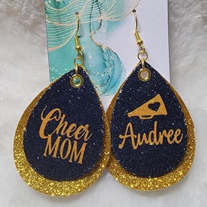 May include: A pair of gold and navy blue glitter earrings. One earring has the text "Cheer Mom" and the other earring has the text "Audree" and a gold megaphone with a heart.