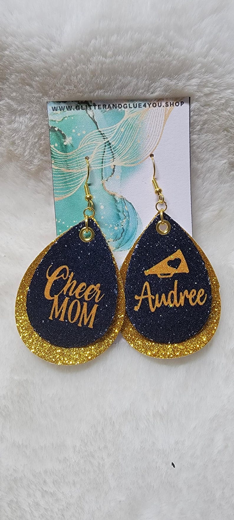 May include: A pair of gold and navy blue glitter earrings. One earring has the text "Cheer Mom" and the other earring has the text "Audree" and a gold megaphone with a heart.