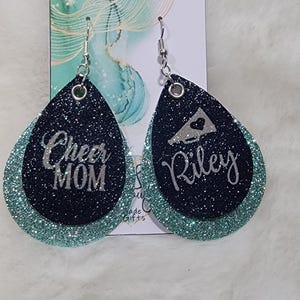 May include: A pair of teal and black glitter earrings with silver lettering. One earring says "Cheer Mom" and the other says "Riley" with a silver megaphone graphic.