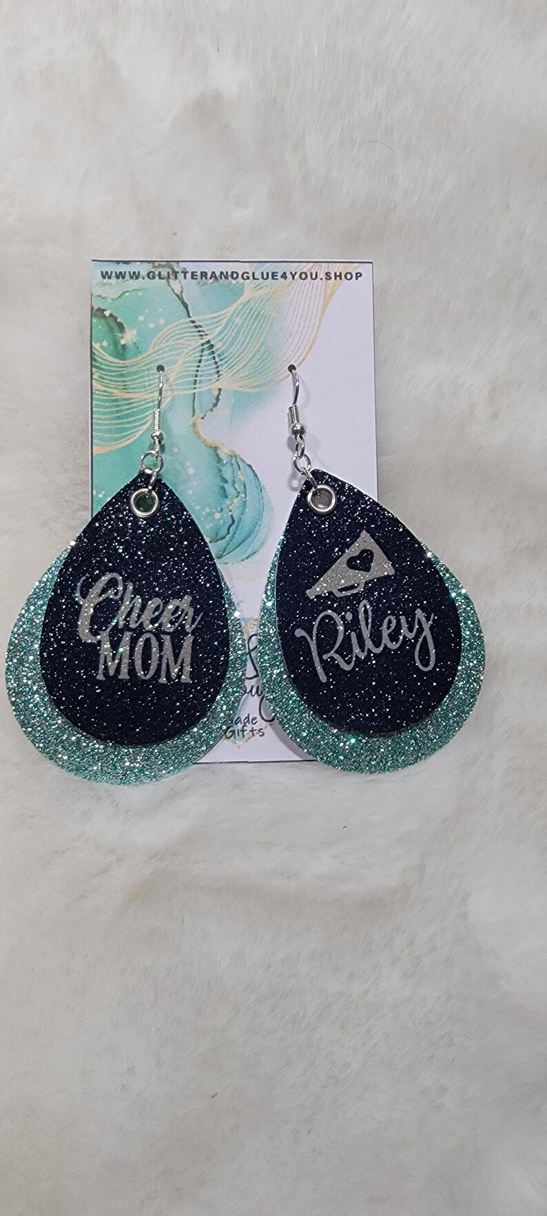 May include: A pair of teal and black glitter earrings with silver lettering. One earring says "Cheer Mom" and the other says "Riley" with a silver megaphone graphic.