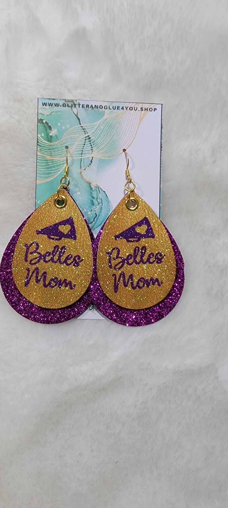 May include: A pair of gold and purple glitter earrings with a purple megaphone and the text "Belles Mom" on each earring.