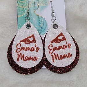 May include: A pair of red and white glitter earrings with a white megaphone and the text "Emma's Mama" in red glitter.