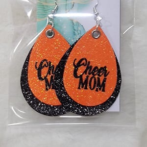 May include: A pair of orange and black glitter earrings with the text "Cheer Mom" on one earring and "Cheer" on the other earring.