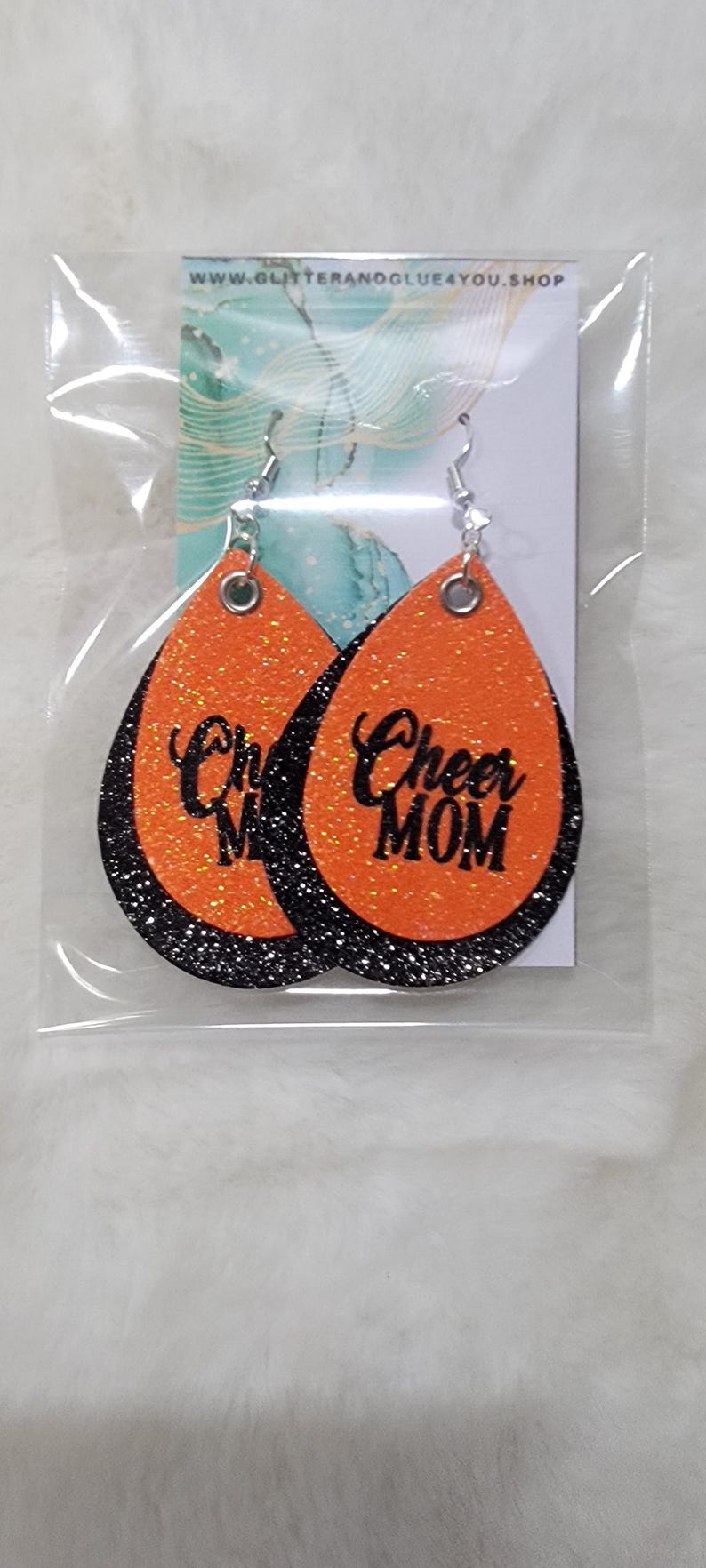 May include: A pair of orange and black glitter earrings with the text "Cheer Mom" on one earring and "Cheer" on the other earring.