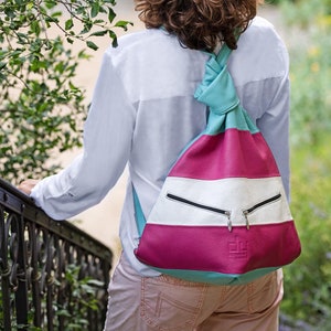 May include: A colorful, striped backpack in teal, white, and magenta. The bag has two zippered pockets and a knotted strap. The person in the image is wearing a white shirt and tan pants.