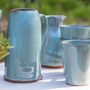 Puede incluir: Un juego de tazas de cerámica azul claro y una jarra. Las tazas están apiladas una encima de la otra y la jarra está en el fondo. Las tazas y la jarra tienen un aspecto rústico y hecho a mano.