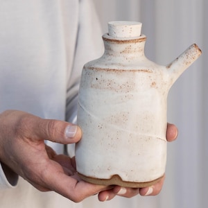 Puede incluir: Una botella de cerámica blanca con un tapón de corcho y un pico. La botella tiene un esmalte marrón moteado y una superficie ligeramente texturizada.