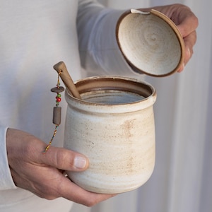 Könnte beinhalten: Eine weiße Keramikdose mit braunem Deckel und einem Holzlöffel, der an einer Schnur mit bunten Perlen befestigt ist. Die Dose ist mit braunen Flecken versehen und hat einen braunen Rand.