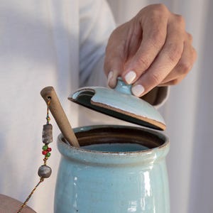 Könnte beinhalten: Eine hellblaue Keramikdose mit Deckel und einem Holzlöffel. Die Dose hat ein rustikales, handgemachtes Aussehen und ist perfekt zum Aufbewahren von Honig, Marmelade oder anderen Vorratsartikeln.