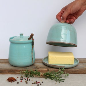 May include: A light blue ceramic butter dish with a lid and a wooden spreader. The dish is filled with a stick of butter and is sitting on a wooden surface.