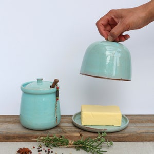 Peut inclure: Un beurrier en céramique bleu clair avec un couvercle et un couteau à beurre en bois. Le beurrier est posé sur une planche en bois avec un morceau de beurre, un brin de romarin et des épices.