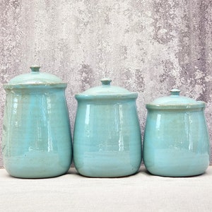 Puede incluir: Conjunto de tres botes de cerámica con tapas en un esmalte azul claro. Los botes son redondos y tienen una base ligeramente acampanada.