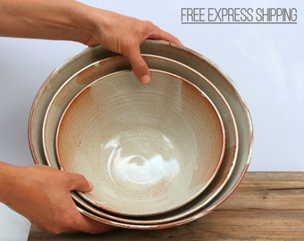 May include: Set of three ceramic bowls in various sizes. The bowls are a light beige color with a brown rim and a subtle swirl pattern.