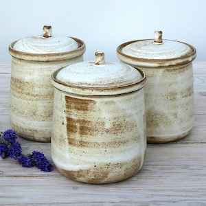 May include: Three white ceramic canisters with brown speckled glaze and lids. The canisters are arranged in a row on a light wood surface.