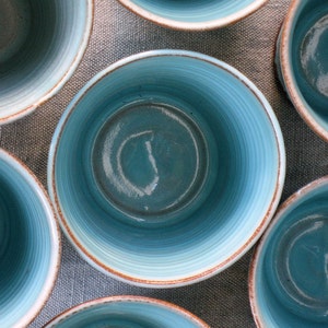 May include: A set of six blue ceramic bowls with a light blue glaze and a white rim. The bowls are arranged in a circle on a textured surface.
