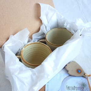 May include: Two handmade ceramic mugs with a light brown glaze and a white interior are nestled in a white tissue paper-lined box. The box is open and the mugs are visible. A small ceramic tag with a blue gemstone is visible in the foreground.