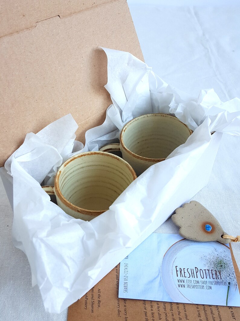 May include: Two handmade ceramic mugs with a light brown glaze and a white interior are nestled in a white tissue paper-lined box. The box is open and the mugs are visible. A small ceramic tag with a blue gemstone is visible in the foreground.