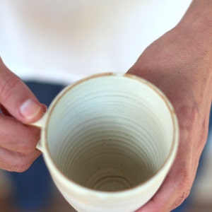 Pu&ograve; includere: Una tazza in ceramica bianca con una glassa testurizzata leggermente biancastra. La tazza ha una forma arrotondata e una bocca larga e aperta.