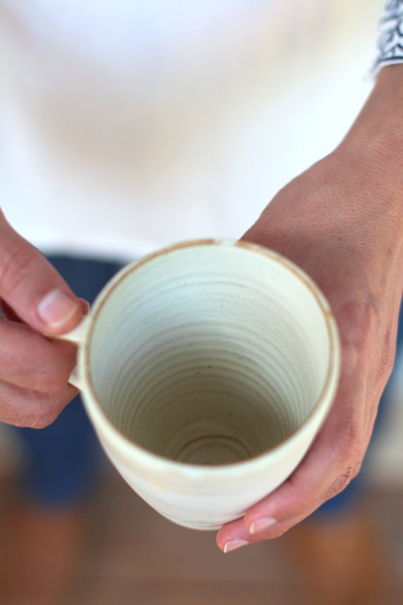 Pu&ograve; includere: Una tazza in ceramica bianca con una glassa testurizzata leggermente biancastra. La tazza ha una forma arrotondata e una bocca larga e aperta.