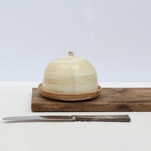 May include: A white ceramic butter dish with a lid and a brown wooden base. A silver butter knife is in front of the dish.