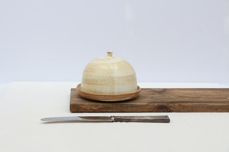 May include: A white ceramic butter dish with a lid and a brown wooden base. A silver butter knife is in front of the dish.