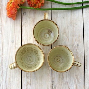 May include: Three ceramic mugs with a light brown glaze and a white interior. The mugs have a textured, swirled design. The mugs are arranged in a triangle shape on a white wooden surface.