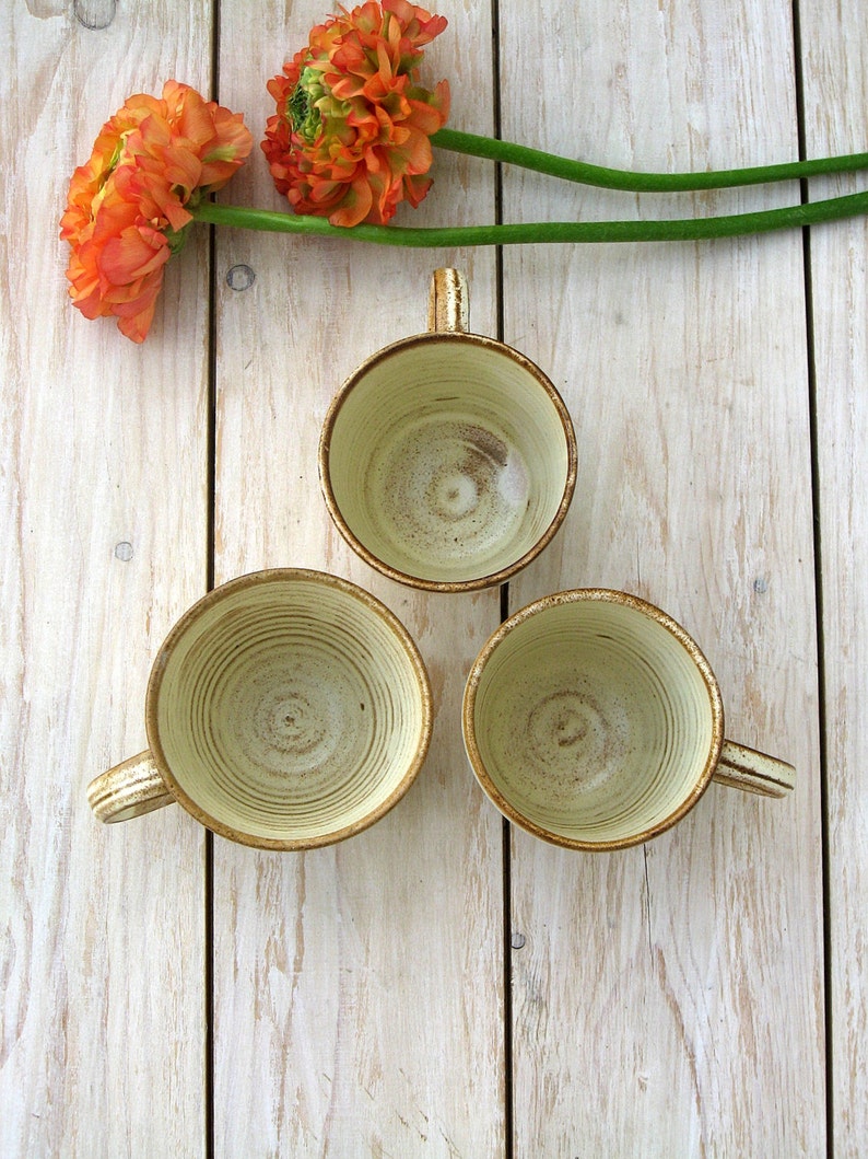 May include: Three ceramic mugs with a light brown glaze and a white interior. The mugs have a textured, swirled design. The mugs are arranged in a triangle shape on a white wooden surface.