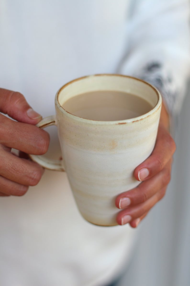 Pu&ograve; includere: Una tazza in ceramica bianca con un bordo beige e una fascia beige intorno al centro, riempita di una bevanda marrone chiaro.