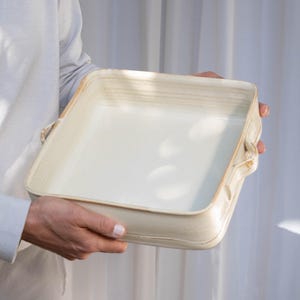 Puede incluir: Un molde para hornear de cerámica blanca con dos asas. El molde es rectangular y tiene un borde ligeramente elevado.