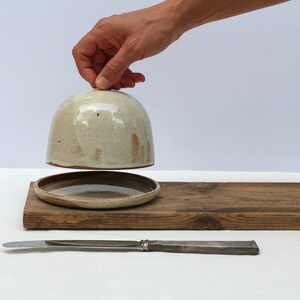 May include: A ceramic butter dish with a lid, a small plate, and a butter knife. The dish is a light beige color with brown speckles. The plate and knife are on a wooden board.