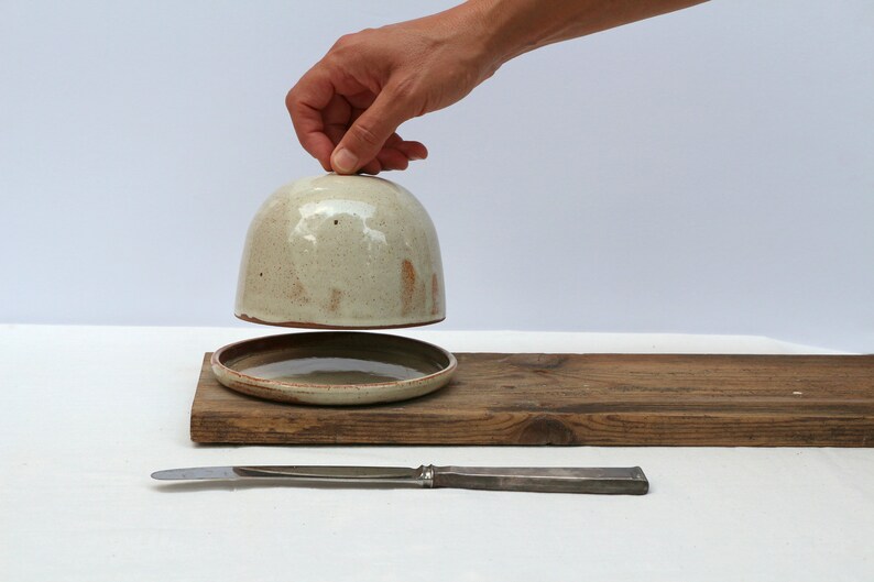 May include: A ceramic butter dish with a lid, a small plate, and a butter knife. The dish is a light beige color with brown speckles. The plate and knife are on a wooden board.
