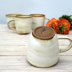 May include: Set of four white ceramic mugs with brown glaze and a rustic look. The mugs have a rounded shape and a handle. The mugs are arranged in a row on a wooden surface.