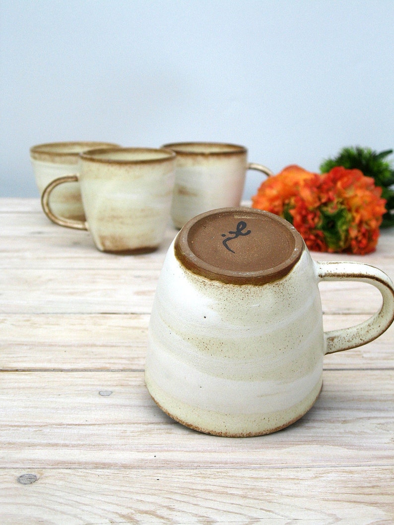 May include: Set of four white ceramic mugs with brown glaze and a rustic look. The mugs have a rounded shape and a handle. The mugs are arranged in a row on a wooden surface.