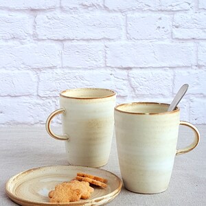 Pu&ograve; includere: Due tazze in ceramica con una glassa bianca e un bordo marrone, una riempita con un liquido marrone chiaro. Le tazze sono appoggiate su una tovaglia di lino beige accanto a un piccolo piatto di ceramica bianco con tre biscotti marroni.