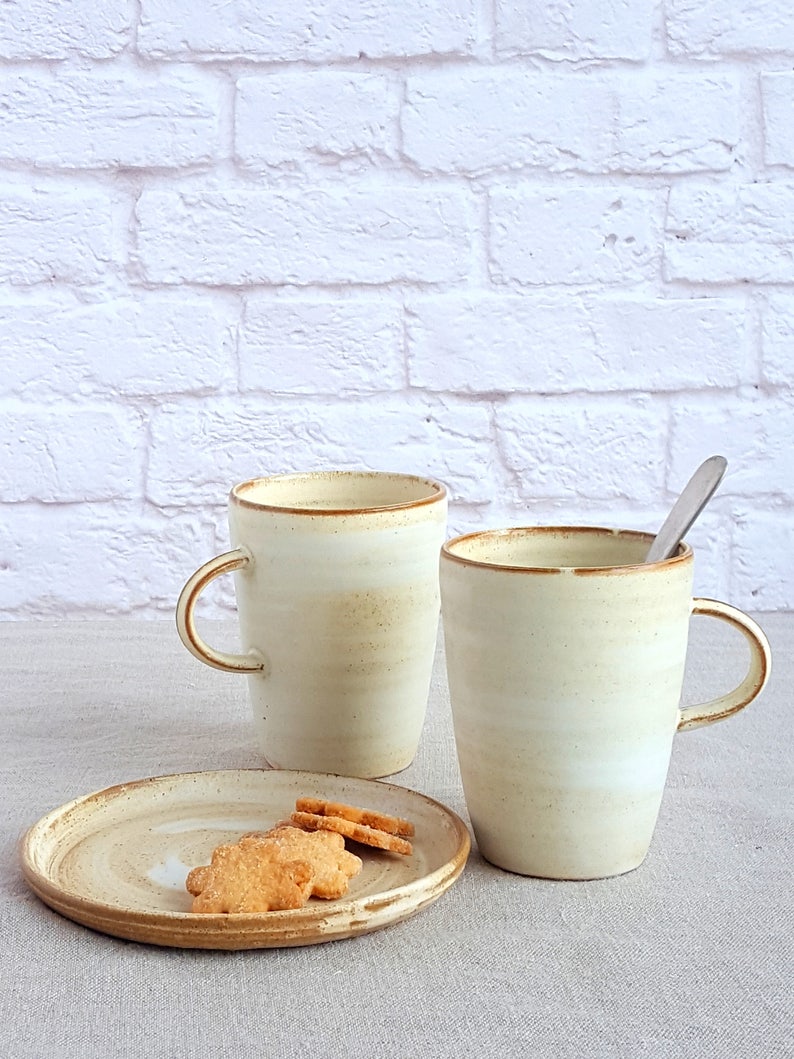 Pu&ograve; includere: Due tazze in ceramica con una glassa bianca e un bordo marrone, una riempita con un liquido marrone chiaro. Le tazze sono appoggiate su una tovaglia di lino beige accanto a un piccolo piatto di ceramica bianco con tre biscotti marroni.