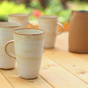 Pu&ograve; includere: Quattro tazze in ceramica bianca con bordi marroni e un'anfora in ceramica marrone su una superficie in legno. Le tazze hanno una smaltatura marrone chiaro con una striscia bianca.