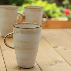Pu&ograve; includere: Tre tazze in ceramica bianca con bordi marroni e una glassa macchiettata. Le tazze sono su un tavolo di legno.