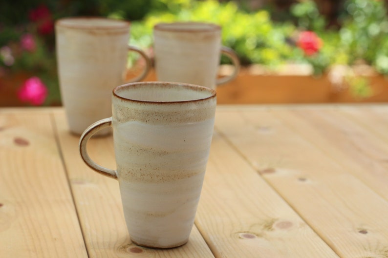 Pu&ograve; includere: Tre tazze in ceramica bianca con bordi marroni e una glassa macchiettata. Le tazze sono su un tavolo di legno.
