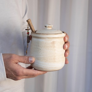 May include: A white ceramic jar with a lid and a wooden spoon. The jar has a rustic, handmade look and is perfect for storing honey or other condiments.