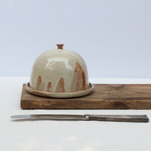 May include: A white ceramic butter dish with a brown glaze and a dome lid. The dish is sitting on a wooden board with a silver butter knife.